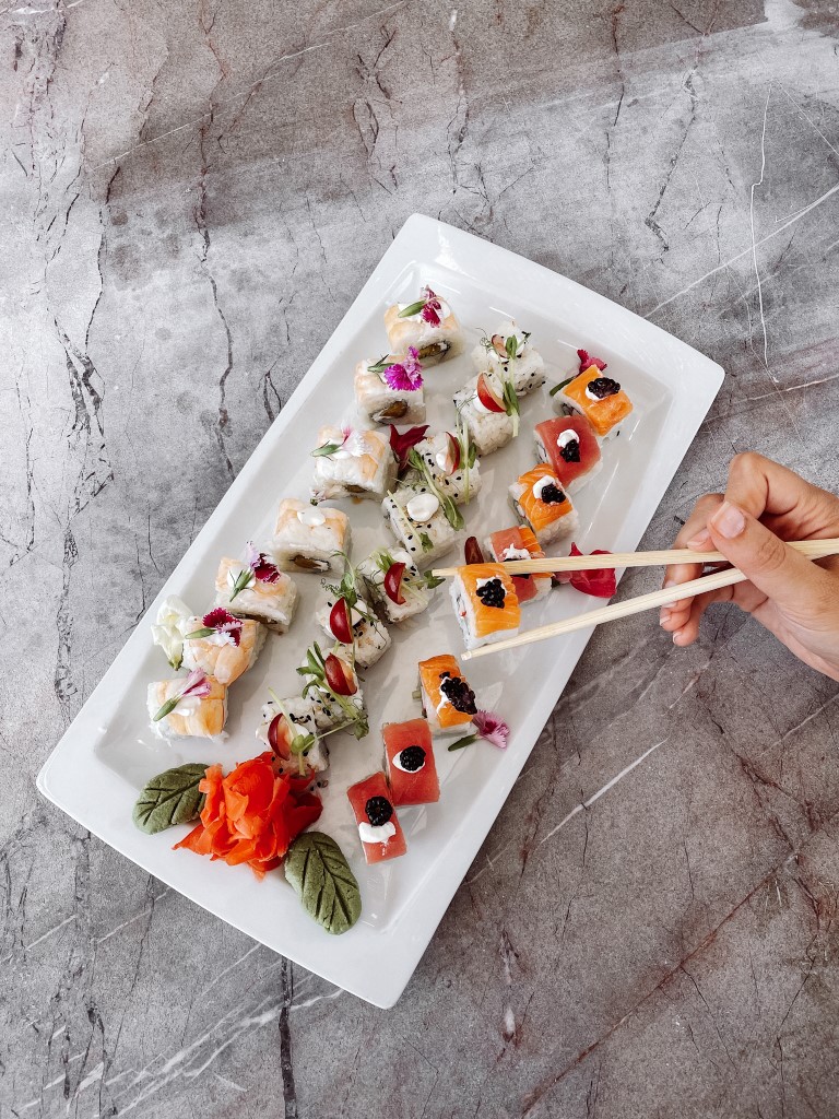 A plate of sushi with three rows of rolled sushi and a hand holding a pair of chopsticks next to it 