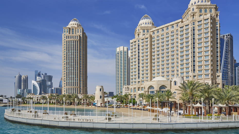 A large, majestic hotel building next to the sea, with modern skyscrapers seen in the distance