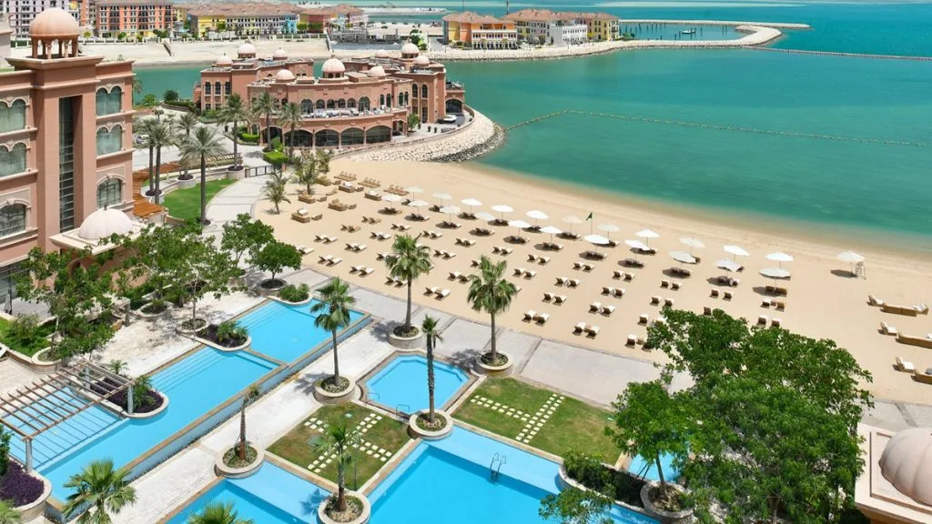 The exterior of a beachfront resort with several pools lined by palm trees, a beach area with sunbeds and umbrellas, and the sea 