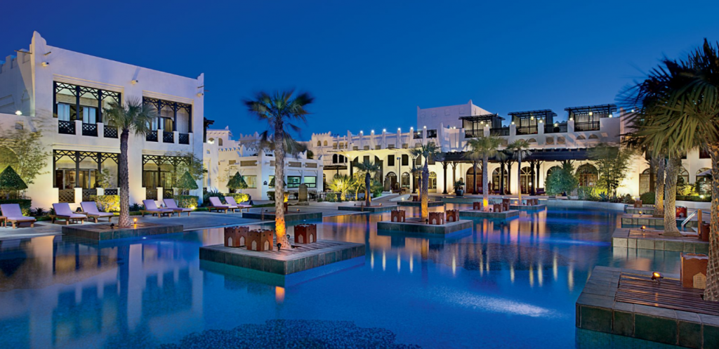 A large pool with palm trees in the center, surrounded by white buildings at dusk