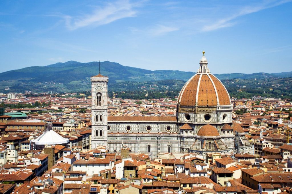 Image of Santa Maria dei Fiori with Brunelleschi's Dome and Giotto's Bell Tower in the center. The building is surrounded by terracota rooftops, and green hills in the background. It's one of the best views of Florence!