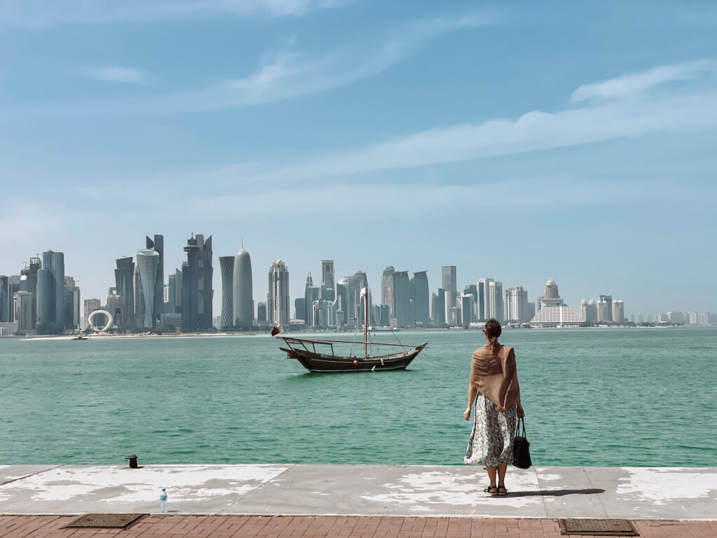 A woman standing in Doha Corniche, overlooking the sea, a traditional dhow boat, and skyscrapers in the background