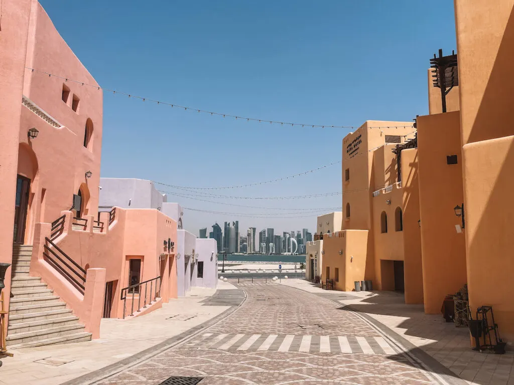 A broad road lined by vibrantly painted buildings, leading up to Doha Corniche. Skyscrapers are seen in the distance