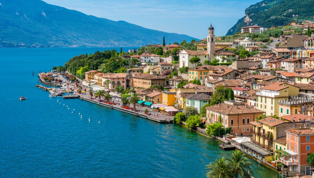 AN image of Lake Garda, with blue waters on the left, picturesque houses on the shoreline, and mountains in the background