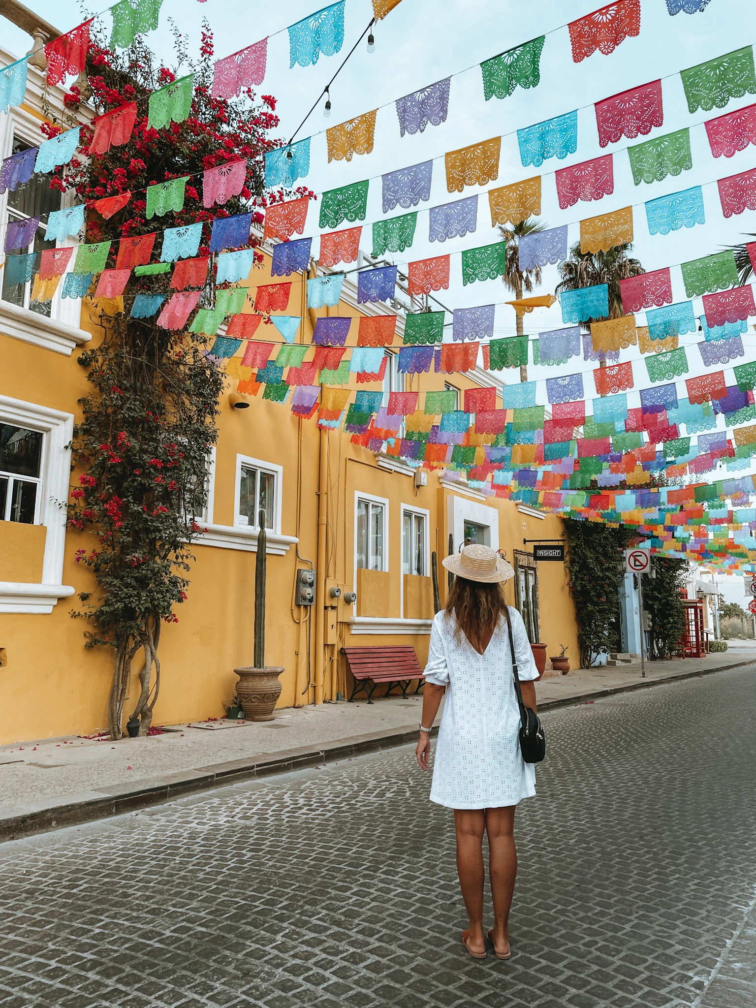 A woman in a white dress and a hat, standing under the colorful flags of a cobblestone street in Los Cabos