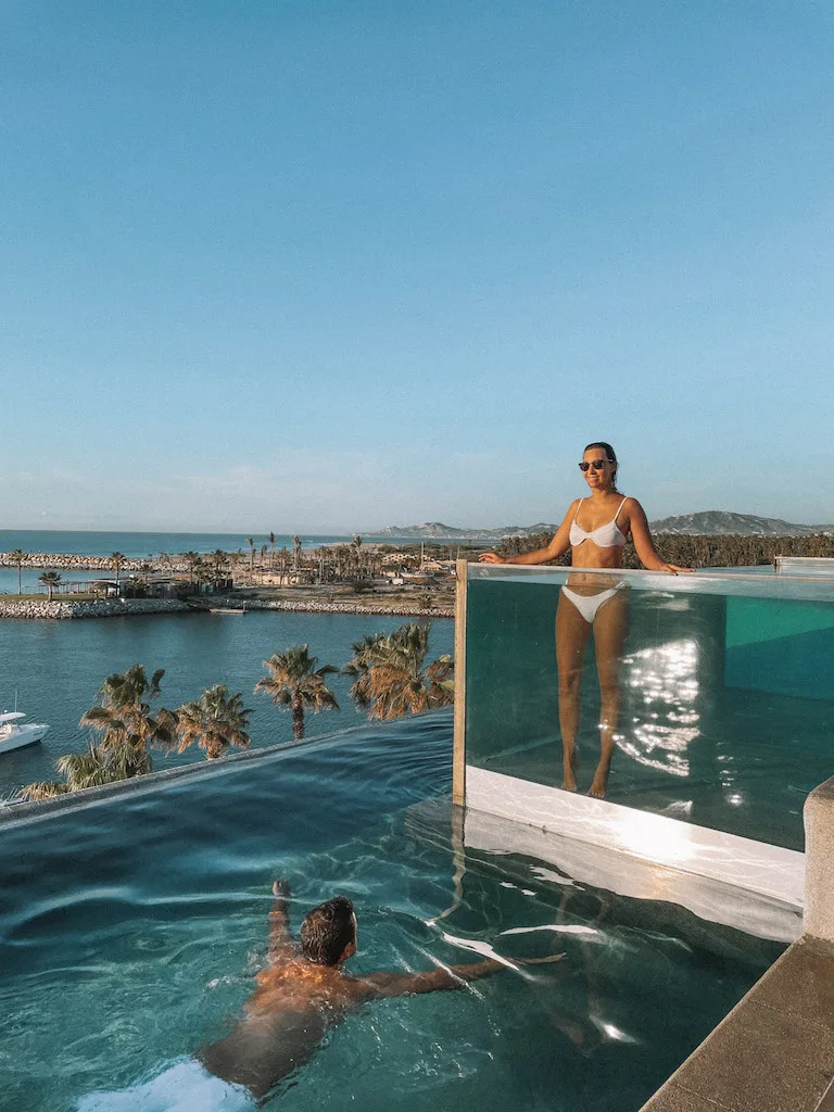 A woman standing in a glass pool, and a man swimming  in the swimming pool below 