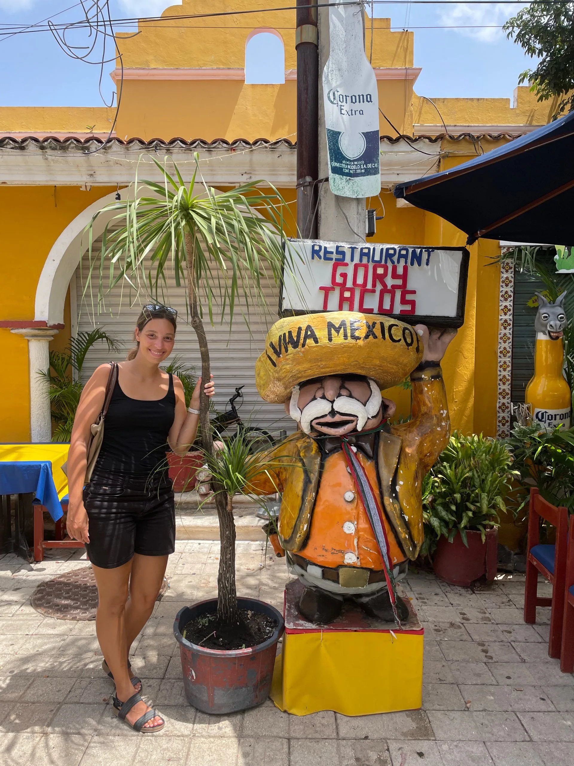 A woman smiling next to a sculpture of a Mexican man holding a sign that reads Gory Tacos