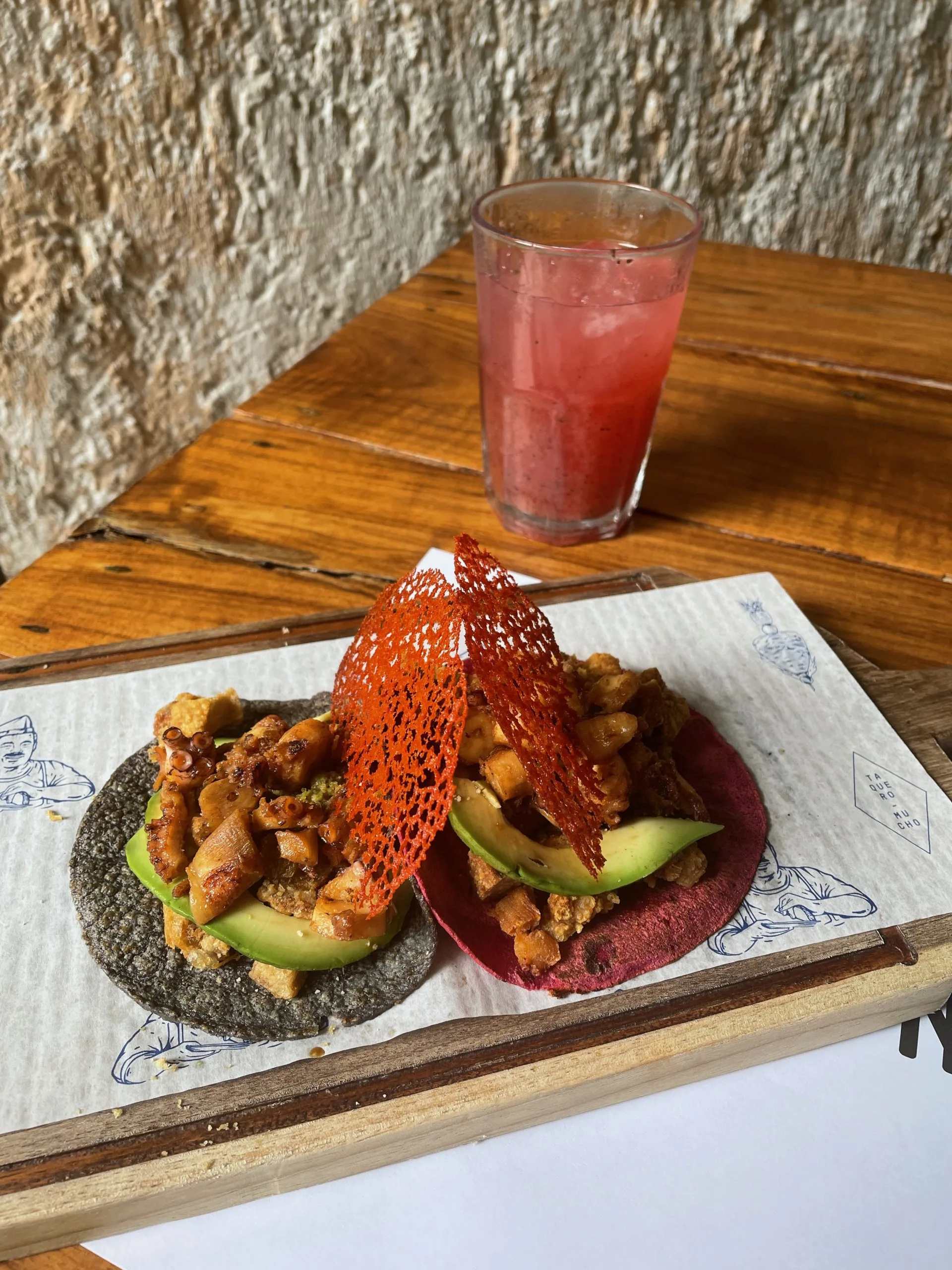 A black and a red tortilla tacos with fillings set on a wooden board, and a glass of juice in the background