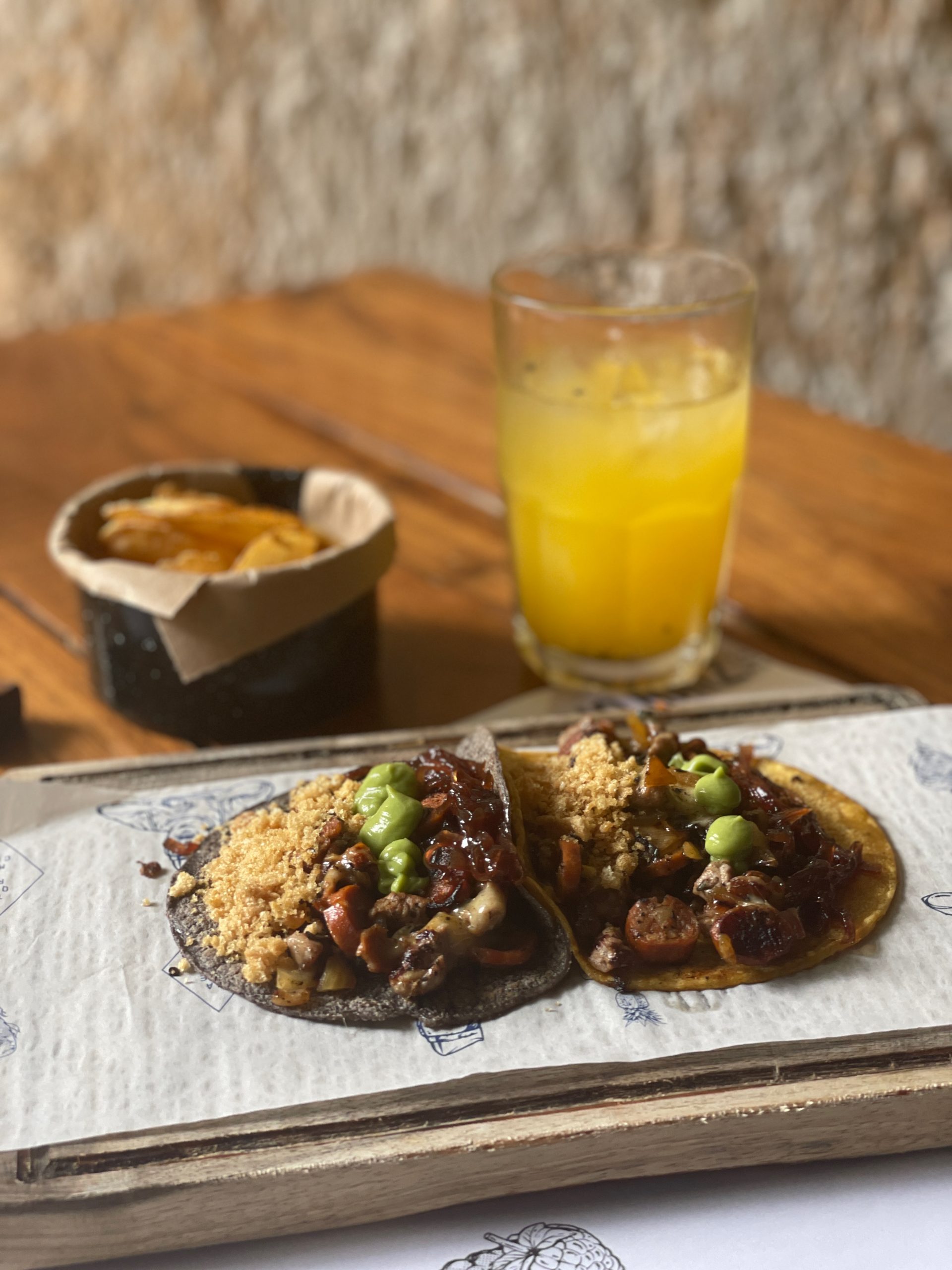 Two tacos served on a wooden board, with a glass of orange juice in the background