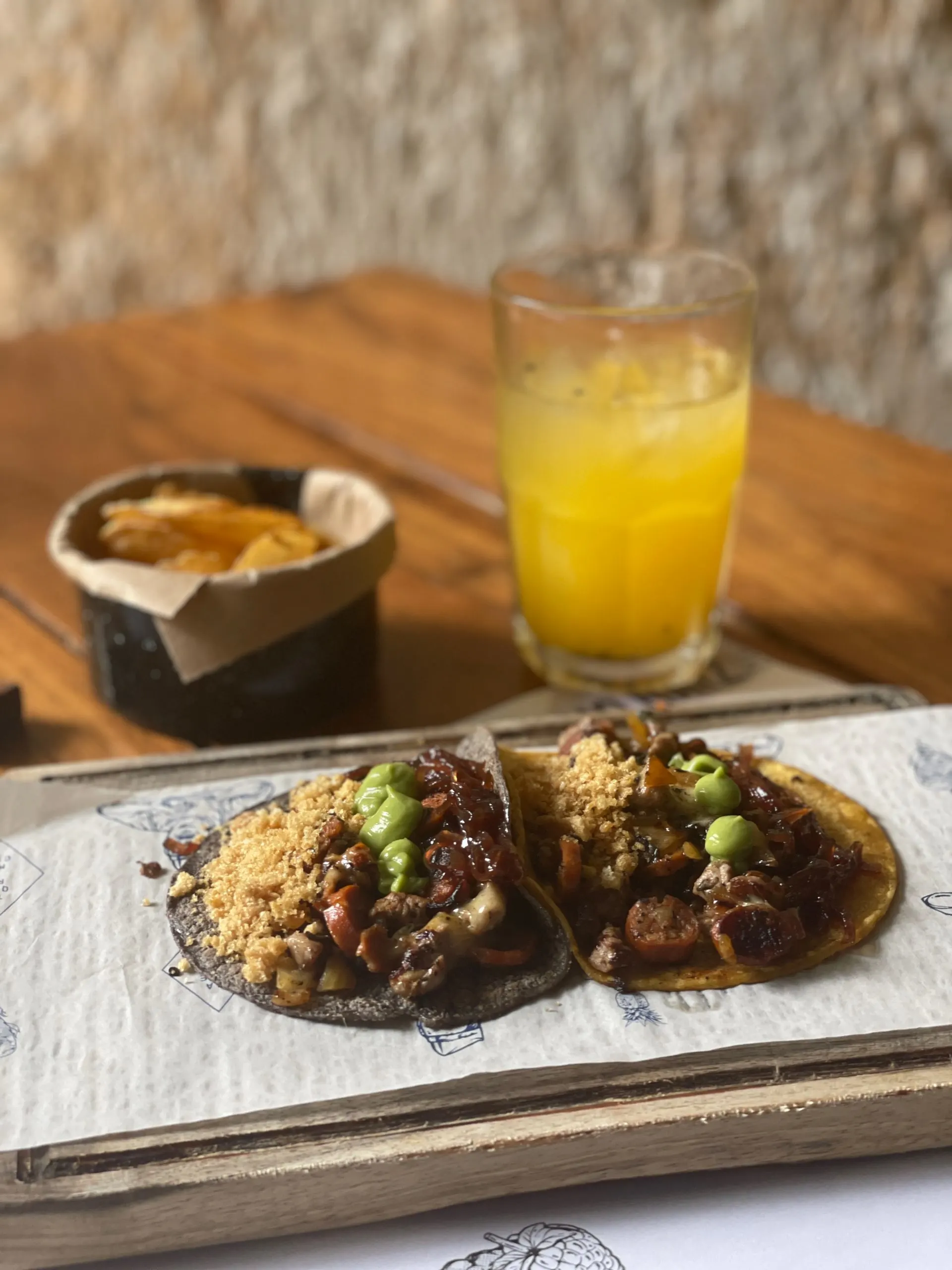 Two tacos served on a wooden board, with a glass of orange juice in the background