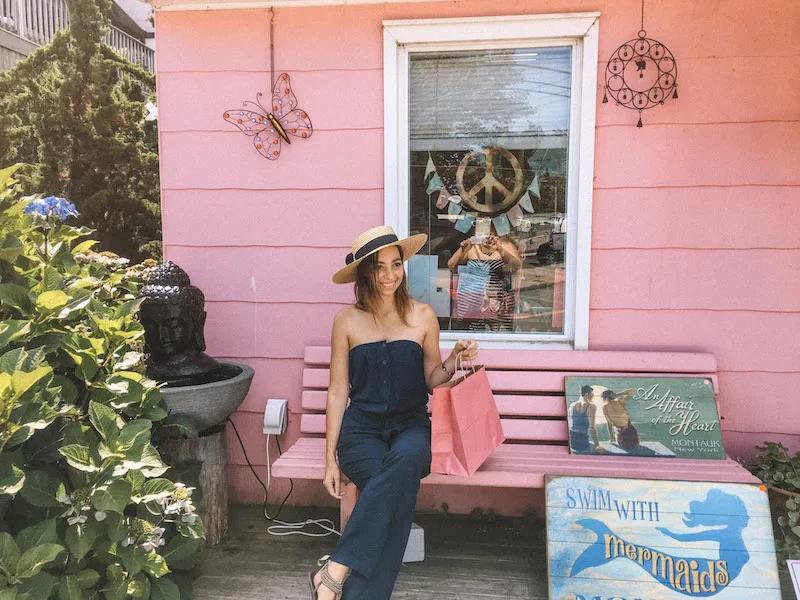 Image of a woman sitting on a pink bench and holding a pink shopping bag, with a pink clothing store behind her in Montauk, inserted in a post about the best things to do in Long Island.  