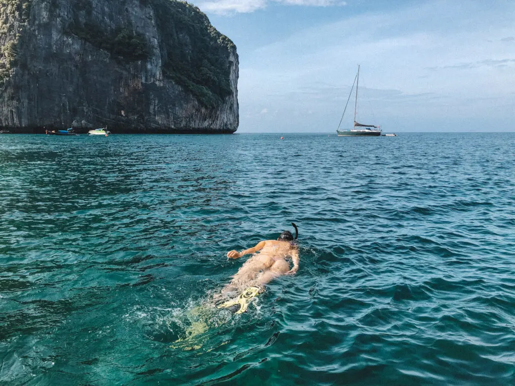 Woman snorkeling in the water, a rock and a sailing boat in the background.