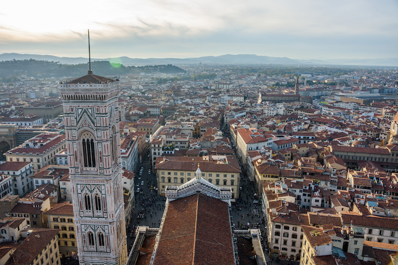 Image of terracota-colored rooftops seen from above, hills in the distance, and a bell tower to the left of the picture, viewed from Brunelleschi's Dome, one of the best views of Florence. 