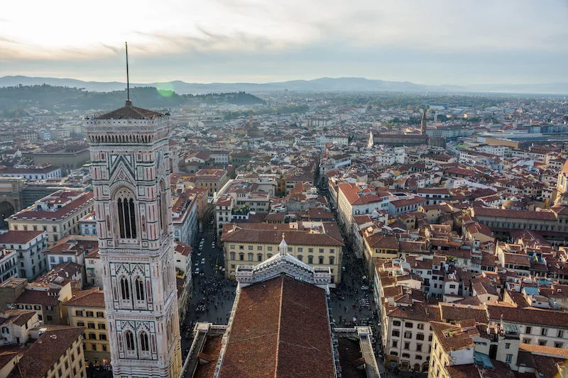 Image of terracota-colored rooftops seen from above, hills in the distance, and a bell tower to the left of the picture, viewed from Brunelleschi's Dome, one of the best views of Florence. 