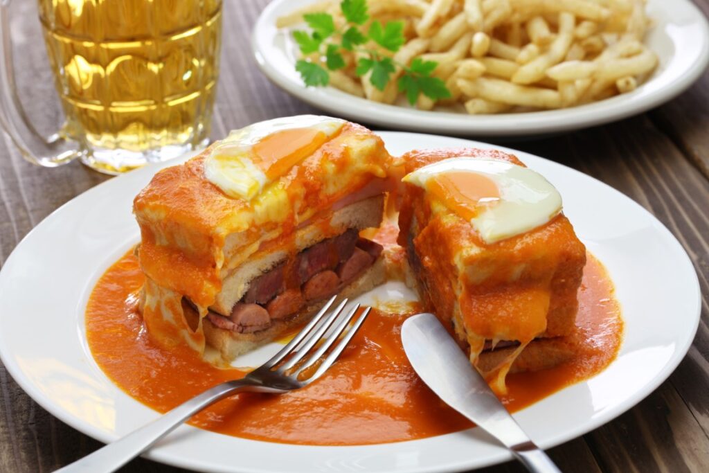 Image of a franchesinha, Porto's traditional dish, with a dish of fries and a glass of beer in the background