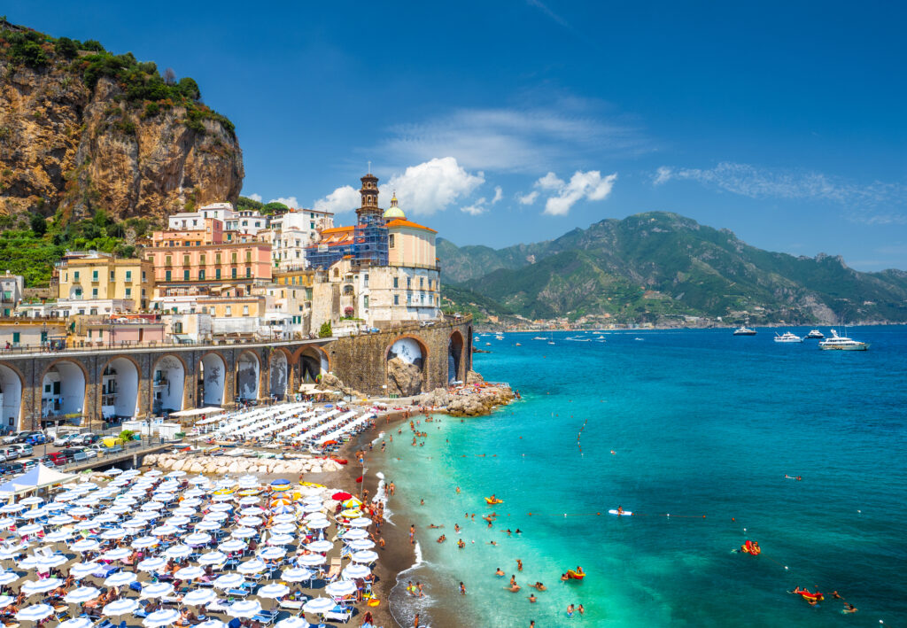 image of one of the best amalfi coast beaches, with the blue sea on the right, cliffs in the background, and a sea of umbrellas and colorful houses on the left