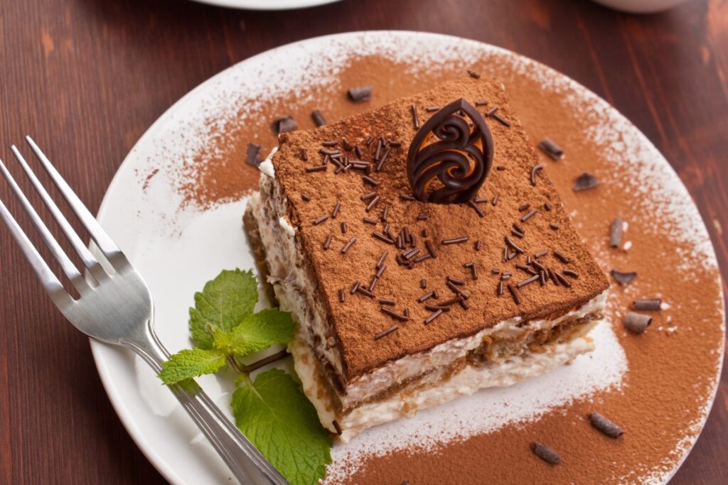 Image of a tiramisu dessert cut in a square shape and  with brown powder on top in a white place with a fork to the side, inserted in a post about the best cooking classes in Naples