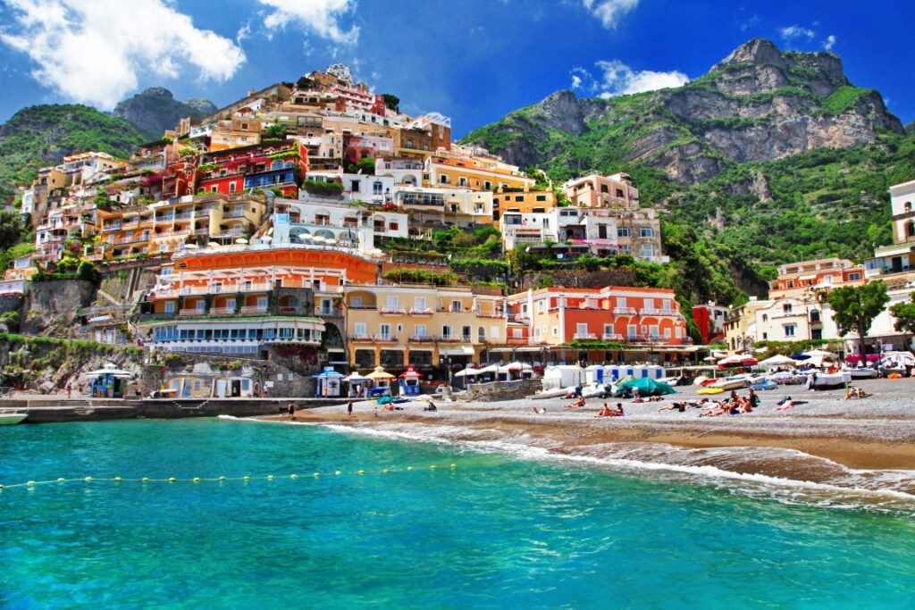 Image of a beach with turquoise waters, and in the background a cliff with colorful houses perched on it, inserted in a post about Sorrento vs Positano vs Amalfi