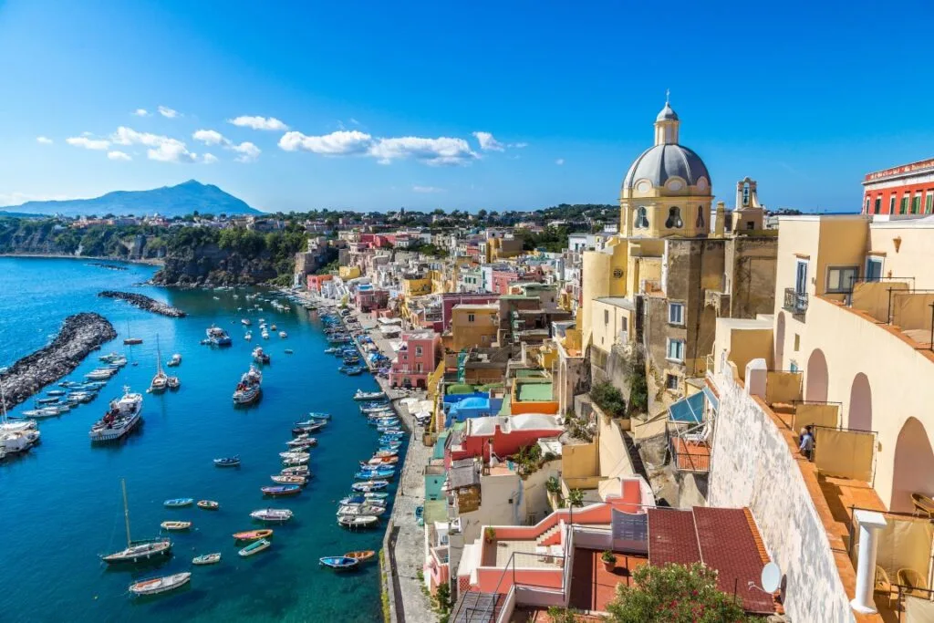 Image of the coastline in Procida, with very colorful houses on the right and the blue sea on the left, dotted with fishing boats. Picture inserted in a post about day trips from Naples