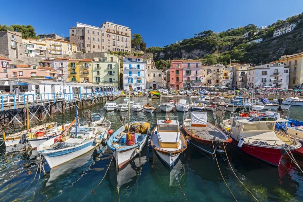 Image of colorful boats on a harbor, with pastel-colored houses in the background and a pier to the left, inserted in a post about Sorrento vs Positano vs Amalfi