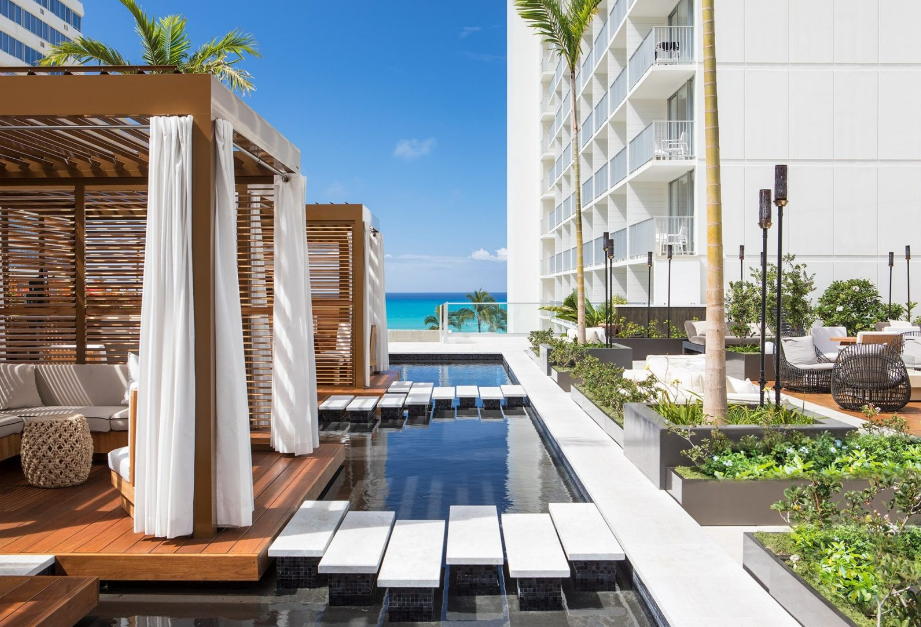 Image of an eco resort in Honolulu, featuring a white building and a patio with furniture on the right, and cabanas on the left, connected by steps above a small pool