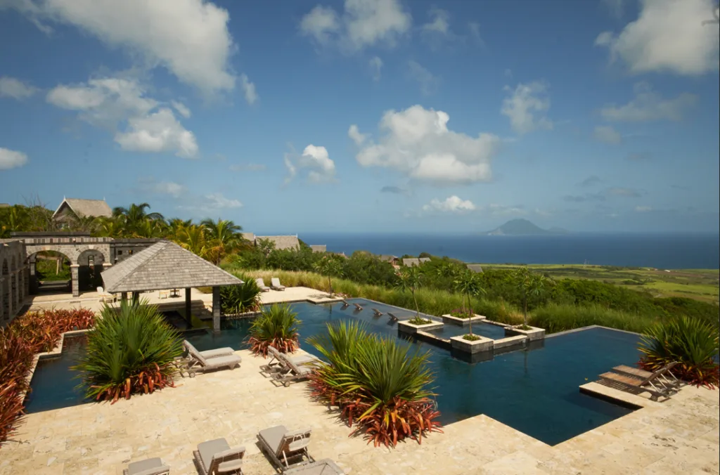 Image of a swimming pool facing green vegetation and the sea, surrounded by sun loungers