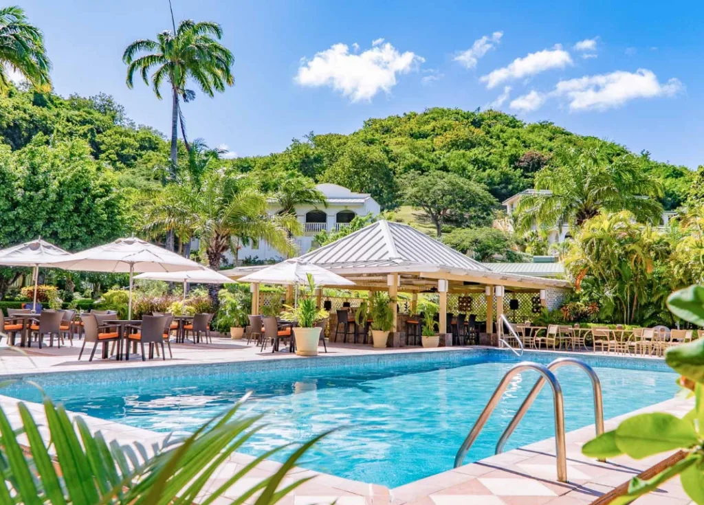 In this picture there's a swimming pool in the foreground, and several outdoor tables with chairs and umbrellas next to it. Lush greenery takes the background of the image