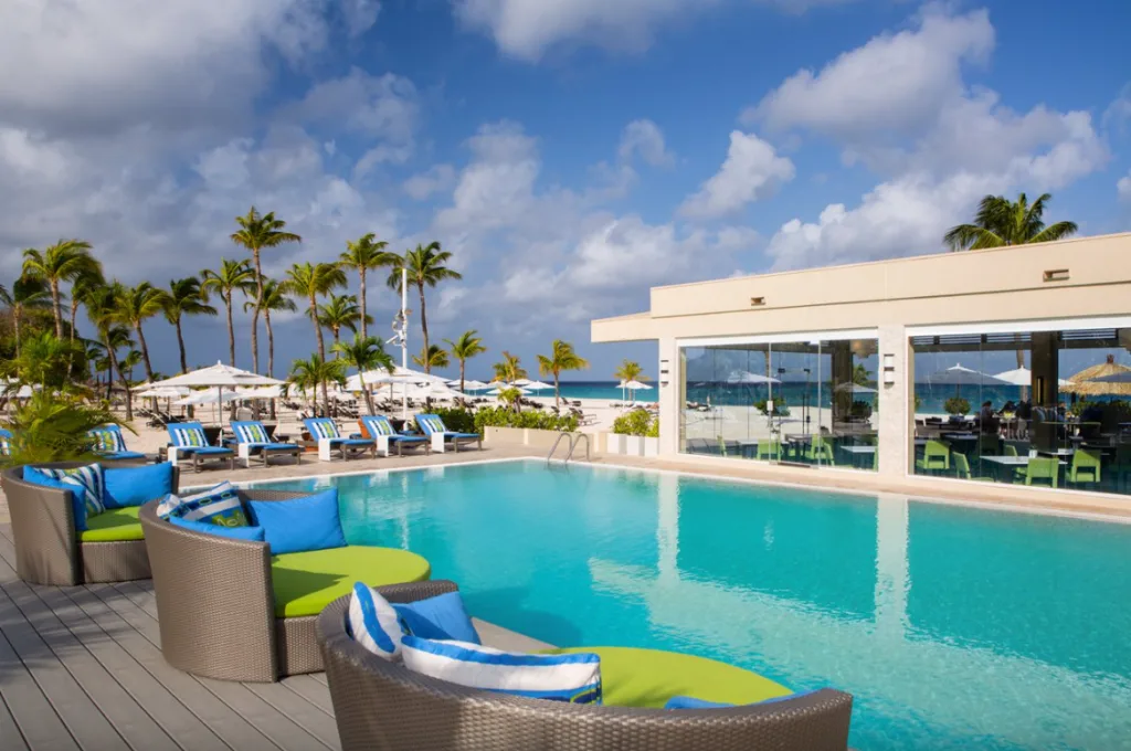 Image of a large swimming pool surrounded by green and blue sunbeds and chairs, a building to the side and palm trees in the background