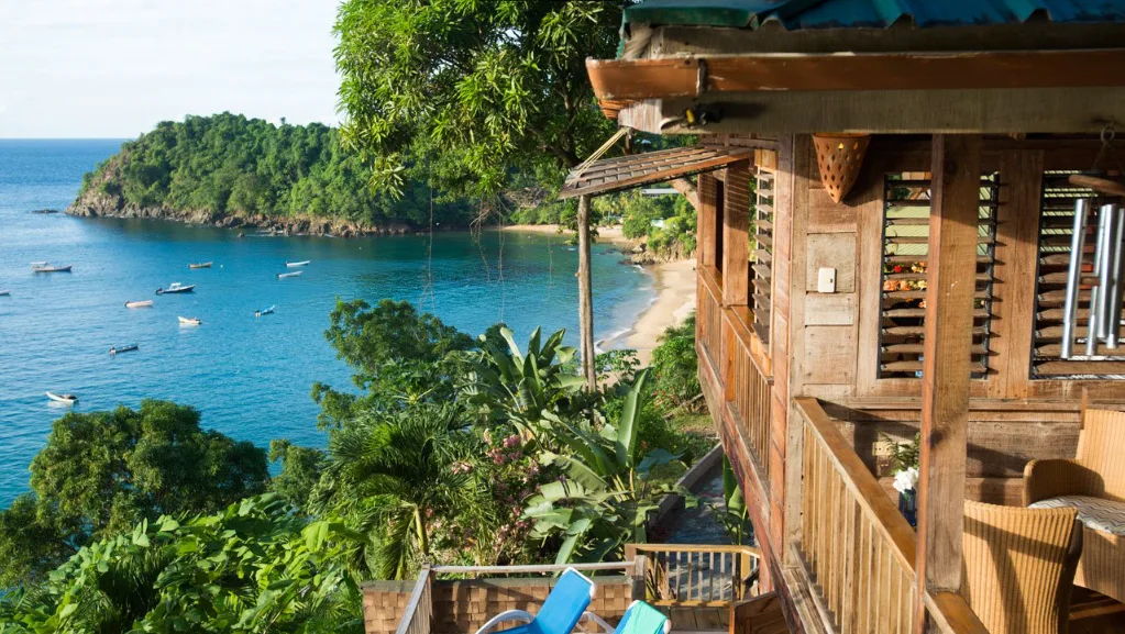 On the right of the image there's a wooden cabin, and on the left the Caribbean Sea with fishing boats, fringed by the sand and lush vegetation