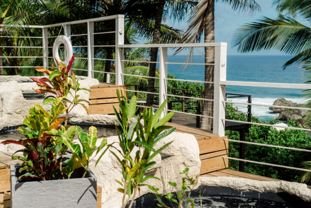 Image of an eco hotel in Barbador, depicting the Caribbean Sea in the background, and the boarwalk to a room surrounded by vegetation