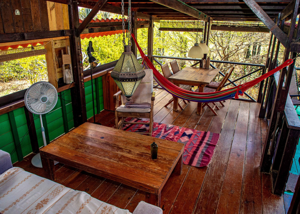 Image of a cabin's dining room, equipped with wooden chairs and a table, a standalone fan, a rug, and a hammock 