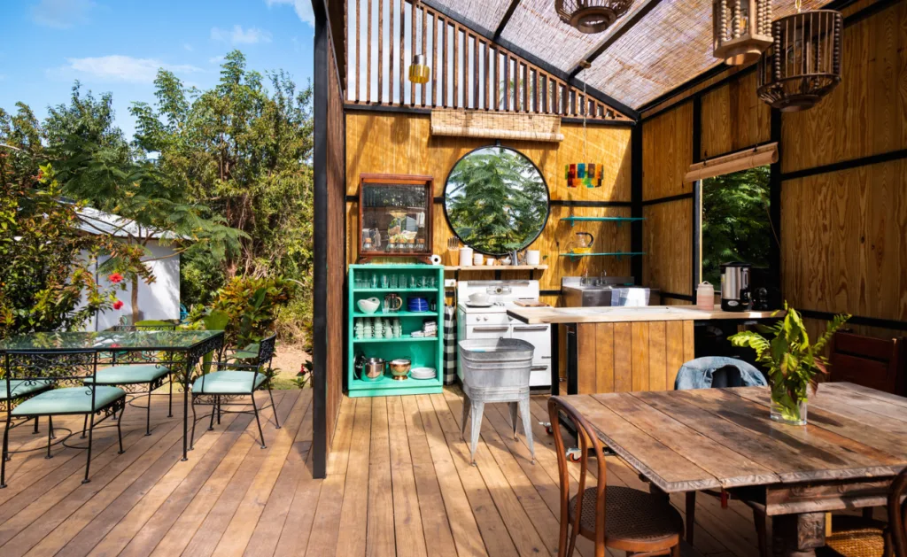 Image of a wooden, open-air room with a table and chairs, and a kitchenette with a bar. On the left side of the image there's outdoor dining furniture and trees 