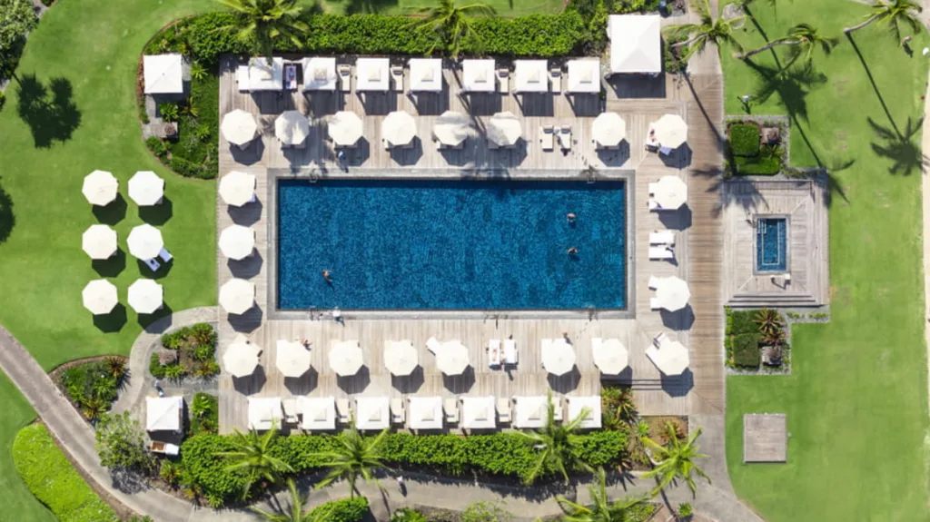 A large swimming pool seen from above, completely surrounded by a deck with white umbrellas, and vividly green grass all around it, with a few palm trees on the sides