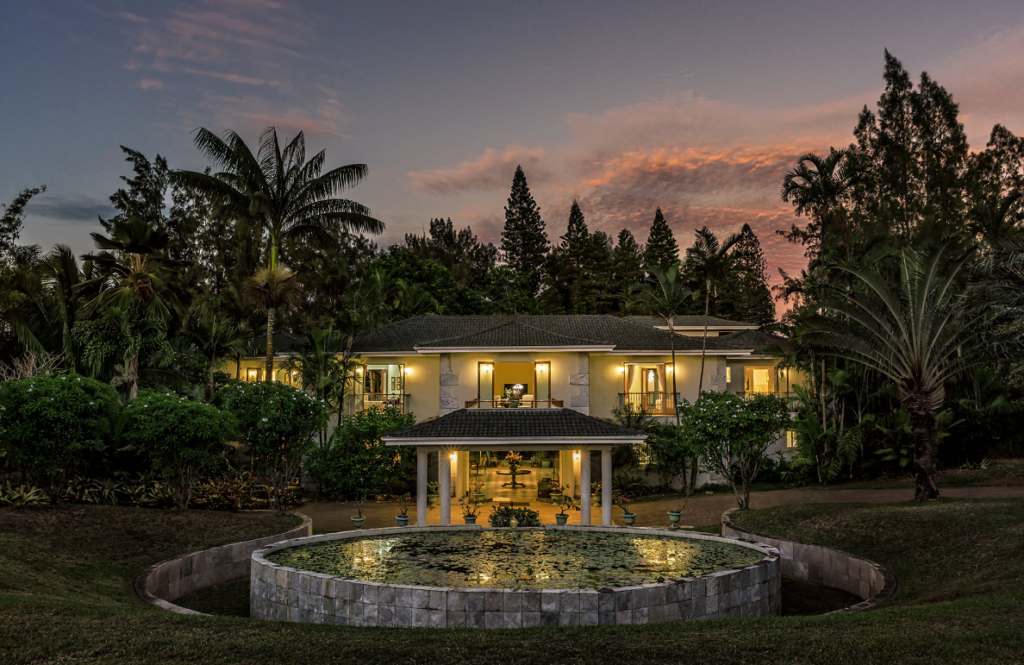 Image of a house in the evening, with all its lights turned on and surrounded by lush foliage. In the foreground there's a circular pong 