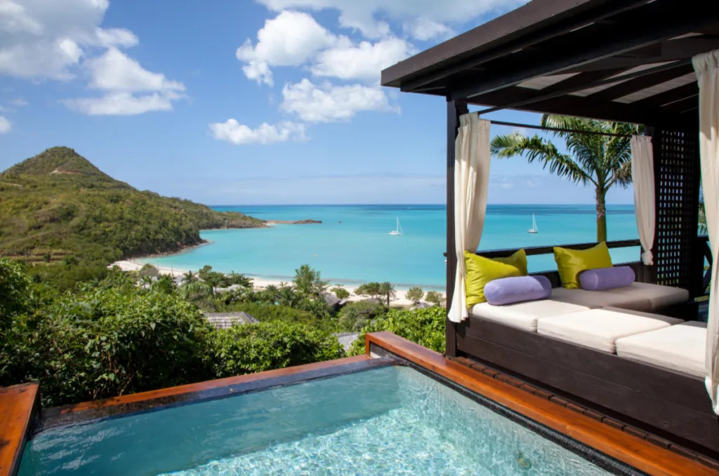 Image of a swimming pool next to a gazebo with cushions, and in the background the Caribbean Sea with a couple of boats and green vegetation. 