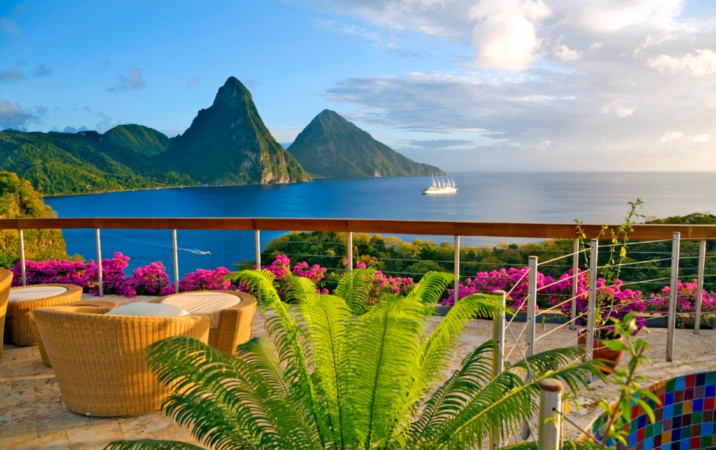 An idyllic image of a terrace with plush outdoor chairs, flowers on the parapet, and the blue ocean in the background, with a boat sailing in the middle. There are tall hills covered in vegetation, and at the forefront of the picture a fern. 