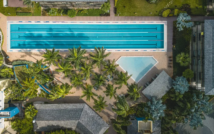 A large swimming pool seen from above is at the top of the image, and below there are palm trees, a building, and a smaller pool