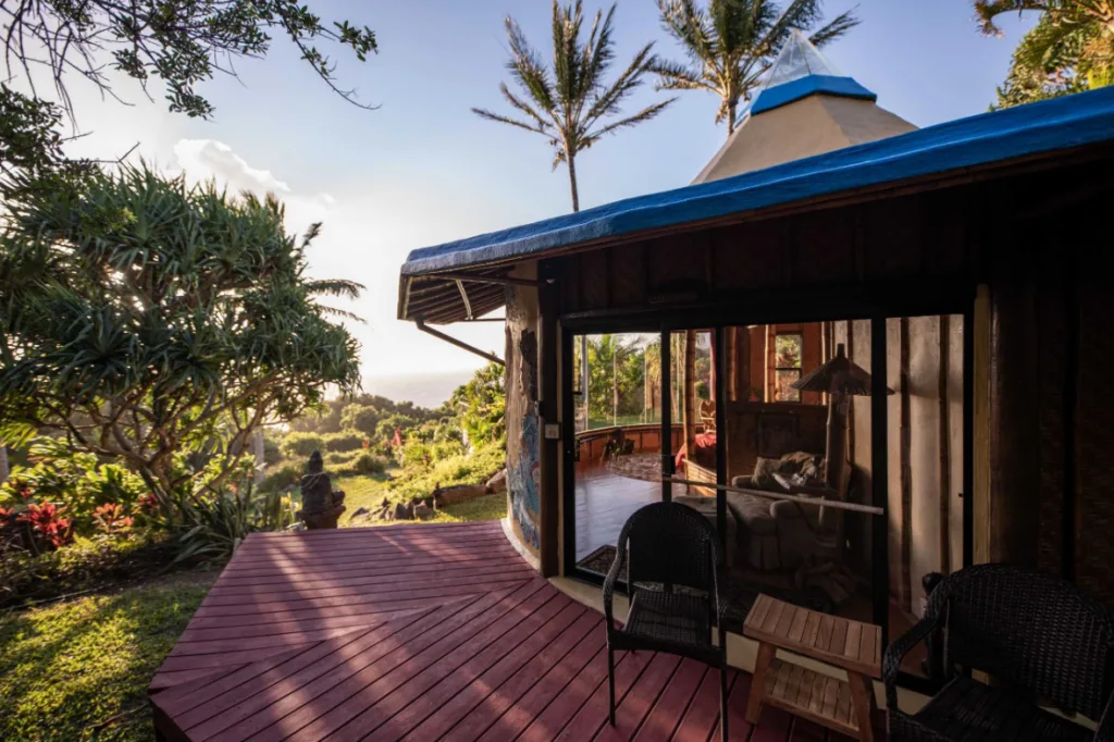 Image of a cabin with a wooden deck and two outdoor chairs, and views of lush gardens