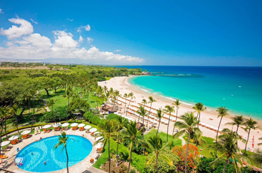 On the right side of the image there's the turquoise ocean, and white sand with orange sun loungers. On the left there are palm trees and a circular swimming pool surrounded by loungers