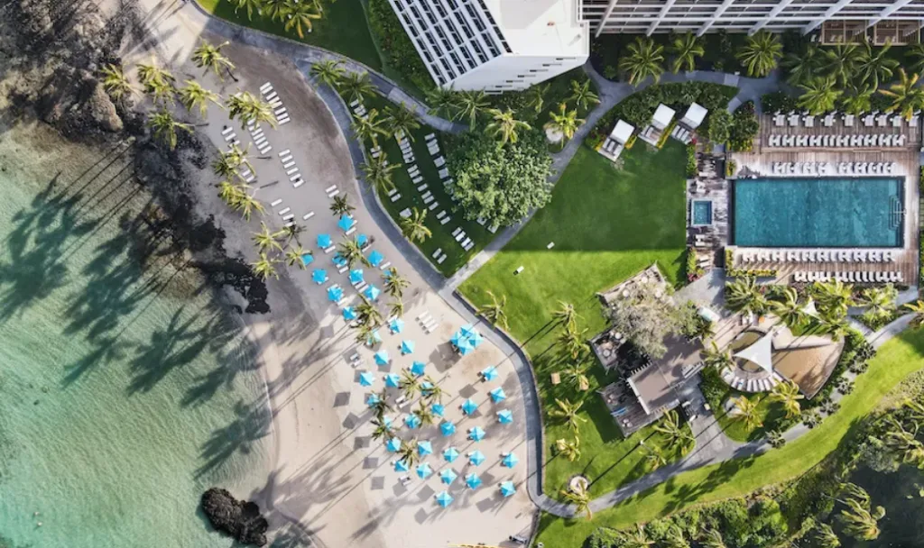 Image taken from above of a resort's recreation area. On the right there's a swimming pool surrounded by loungers, and green areas with a few buildings, and on the left there's the beach with blue umbrellas, palm trees and green waters