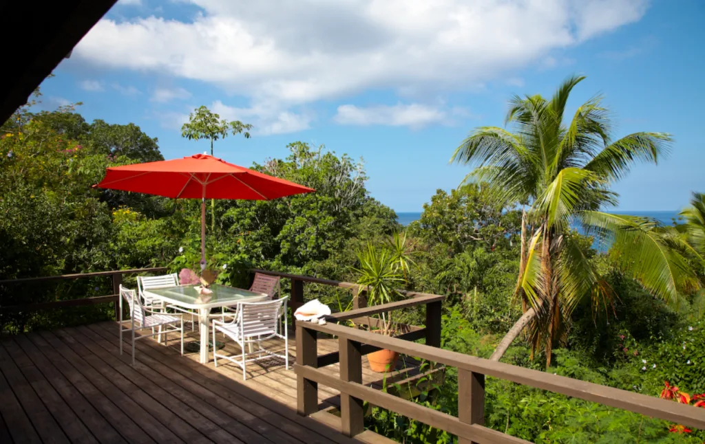 Image of a wooden deck eqipped with a white outdoor table, four chairs, and an orange umbrella, surrounded by green, luscious vegetation