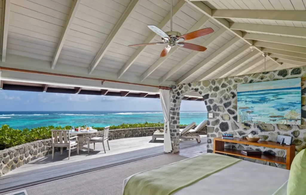 Image of an eco resort in Saint Vincent and the Grenadines, showcasing a double bed in an open-air room that overlooks the blue Caribbean Sea, with a terrace and outdoor dining furniture