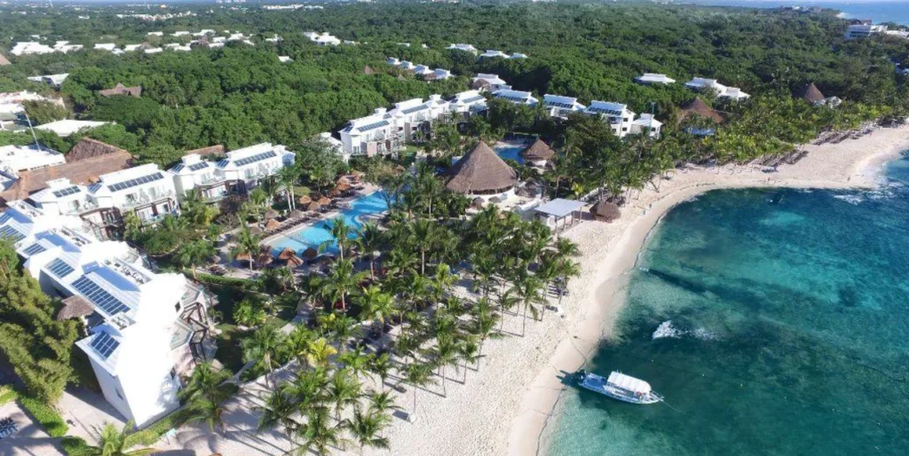 The Caribbean Sea can be seen on the right of the image, with a white yacht on the shore, the white-sand beach dotted with palm trees, and on the left dense vegetation and a large white building with a swimming pool in front of it