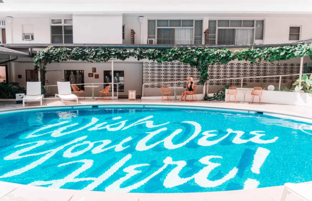 Image of an oval-shaped swimming pool with the letters ''Wish You Were Here'', and a woman in a black swimsuit and a hat sitting on a chair by the pool