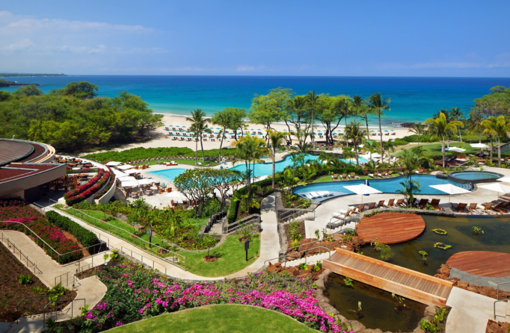 Image of a hotel's recreation area, with two swimming pools, palm trees and green vegetation, a beach area and the ocean in the background, and a pond qith a wooden bridge