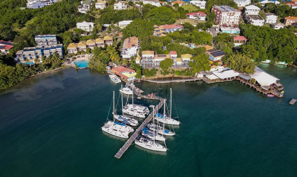 Image taken from above of the sea with nine white boats, and the coastline showcasing green vegetation and dotted with buildings and houses in the island of Grenada