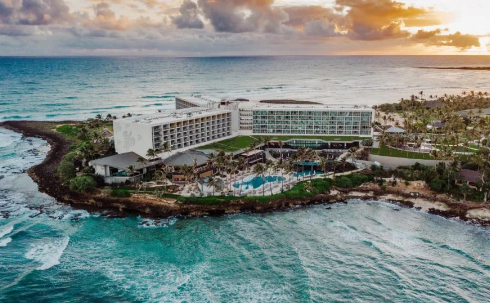 Image of an eco resort in Hawaii, surrounded by the ocean and showcasing two suimming pools and palm trees, seen from a distance