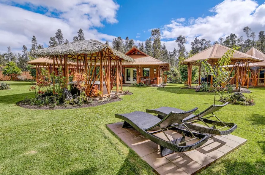 Image of a Hawaii eco resort's cabanas, scattered in a green field with trees and the sky in the background, and two sun loungers in the foreground