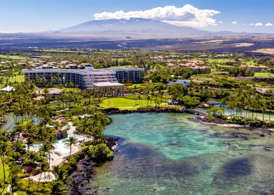 A resort in Hawaii seen from afar, featuring a lagoon in the foreground, a large building in the center, and green fields, palm trees, smalles buildings and pools all around it