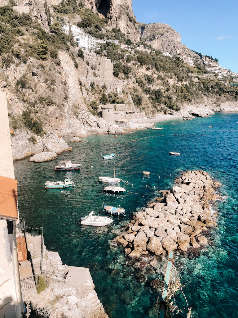 Image of Positano's coastline featuring a  steep cliff covered in vegetation on the left, and the blue Mediterranean Sea dotted with fishing boats on the right